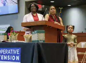 Two women speak at a podium during an event, while a young girl stands beside them. Flowers and boxes labeled "SEED" are on the table in front, with a banner in the background.
