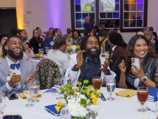 Three smiling guests at a banquet table enthusiastically applauding. The setting features a festive atmosphere with colorful lighting, floral arrangements, and elegantly set tables.