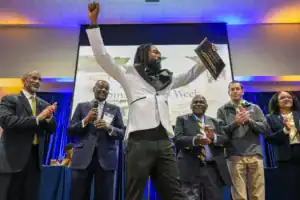 A man in a tuxedo raises a plaque in celebration at an event, surrounded by five applauding individuals. A backdrop displays "Small Business Week.