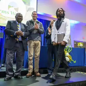 Four people stand on stage at an event, applauding a man in a white tuxedo as he walks past. A blue curtain and flower arrangements decorate the background, with a screen displaying "Farms Week" prominently.