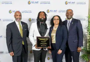 A group of four people pose for a photo, celebrating Darius McKoy, who holds a plaque recognizing him as the 2026 North Carolina Small Farmer of the Year during Small Farms Week.