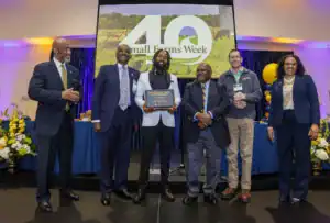 Six people stand together at an event, with one man holding an award. A large screen behind them displays "40 Small Farms Week." The setting features a decorated table and colorful balloons.