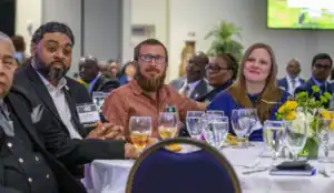 A group of attendees at an event are seated at a round table, enjoying the program. Some are engaged and smiling, with drinks and plates in front of them, amidst a well-decorated setting.