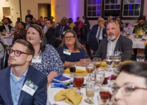 Guests attentively listen at a formal event, seated around tables adorned with glasses and colorful drinks. Some guests wear name tags, and the setting is illuminated with soft lighting.