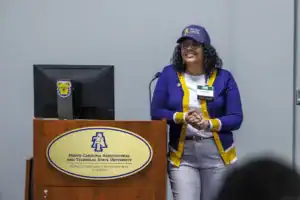 A woman with curly hair, wearing glasses and a blue and yellow cardigan, stands at a podium with a laptop behind her. She is smiling and appears to be speaking at a conference or seminar.