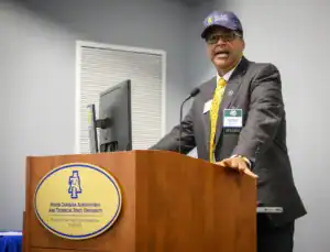 A man in formal attire speaks at a podium with a North Carolina A&T University emblem. He wears a baseball cap and has name tags indicating he is the speaker. There is a computer monitor behind him.
