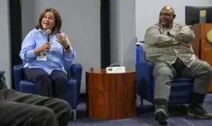 A woman speaking into a microphone sits on a blue chair, expressing herself with hand gestures. A man in a plaid shirt sits nearby with arms crossed, both engaged in a discussion. A wooden table with their names is in front of them.