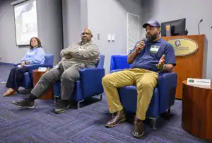 Three speakers sit in blue chairs during a panel discussion. One is speaking into a microphone, while the others listen. A projector screen displays text in the background, and a table with a sign is visible nearby.