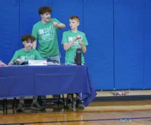 Three young people in green shirts are at a table discussing something. One is pointing, while another listens intently, and a third sits with papers. The background features blue gym walls.