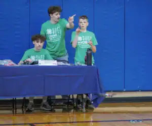 Three students in green shirts interact at a table in a gym. One points while the others observe. A water bottle and competition materials are on the table. Background features blue gym walls.