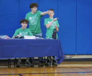 Three young volunteers in green shirts assist with an event at a table, one pointing while another sits with a laptop. A blue gym wall serves as the backdrop.