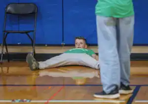 A boy lies on the gym floor with his legs spread out, looking relaxed. A person in light blue pants stands nearby, and a black chair is visible in the background against a blue wall.