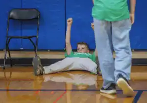 A boy in a green shirt lies on the gym floor with his arms raised in excitement. He is surrounded by other kids in a sports setting, with a chair visible in the background.