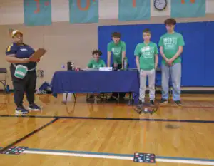 A woman holding a clipboard monitors four students in green shirts as they engage with a drone during an event in a gymnasium, surrounded by tables and equipment.