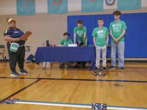 A woman holding a clipboard monitors four students in green shirts as they engage with a drone during an event in a gymnasium, surrounded by tables and equipment.