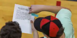 Hands hold a printed sheet detailing a drone mission. The mission involves programming a drone to rotate and activate a 3D scanner in response to environmental needs, with instructions listed.