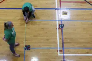 Two people in green shirts are working on a gym floor. One is measuring with a tape while the other observes. Colored lines and markers are laid out on the wooden floor.