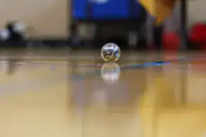 A clear marble rests on a gymnasium floor, reflecting the surrounding environment. The floor has colorful markings and a subtle shine.
