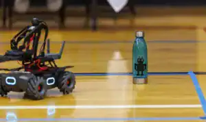 A small robotic vehicle with wheels and antennas is near a teal water bottle featuring a black robot design, set on a wooden gym floor with blue tape markings.