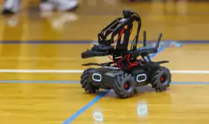 A small robotic vehicle with large wheels and an articulated arm is on a wooden gym floor. It features blue LED lights and wires, showcasing a modern design for robotics or engineering projects.