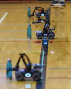 Four small robotic vehicles with articulated arms are lined up on a gym floor, each positioned near a turquoise water bottle, demonstrating a robotics challenge.