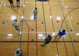 Several people in a gym are working with drones, test equipment on the floor, and taking notes. The gym floor features colored lines and markers for equipment placement.