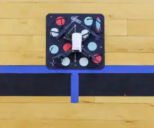 A white drone sits on a colorful mat with circular patterns, positioned on a wooden floor marked with blue tape.