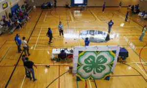 A gymnasium filled with people in blue shirts. Some are gathered at tables, while others participate in activities or watch. A large green and yellow 4-H logo is displayed at the front. Hardwood floors and basketball court markings are visible.