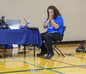A person with curly hair sits anxiously in a chair, covering their mouth with both hands. They are wearing a blue shirt and surrounded by technology, including laptops and a drone, in a gym setting.