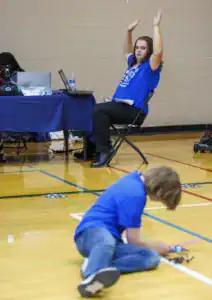 A person in a blue shirt raises their arms while seated at a table with a laptop. In the foreground, a child in a blue shirt is sitting on the floor, focused on a small robotic device.