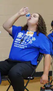 A person with long curly hair sits in a chair, drinking from a water bottle. They wear a blue shirt with a logo and text promoting North Carolina 4-H. A second individual is partially visible in the background.