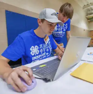 Two young men, one focused on a laptop, engaged in an activity. Both wear blue shirts with inspirational messages. One uses a purple mouse while papers and an envelope are spread on the table.