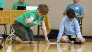 Two kids participate in a robotics activity on a gym floor. One boy kneels, reaching out, while the other sits beside him, focused on a tablet and a clear sphere, with a table in the background.
