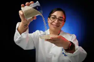A scientist in a lab coat smiles while pouring grains from a beaker into a Petri dish, with a blue and red background. A tattoo is visible on her arm.