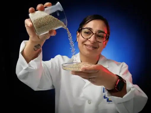 A scientist in a lab coat smiles while pouring grains from a beaker into a Petri dish, with a blue and red background. A tattoo is visible on her arm.