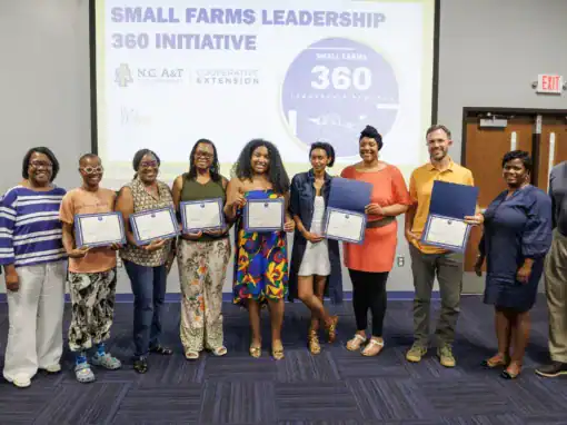 Group of individuals posing with certificates in front of a presentation screen for the Small Farms Leadership 360 Initiative. They are smiling and wearing a variety of clothing styles.