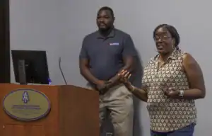 A woman gestures while speaking at a podium, with a monitor visible beside her. A man stands in the background, looking on. Both are engaged in a presentation setting.