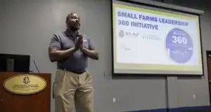 A presenter gestures while discussing the Small Farms Leadership 360 Initiative at a university event. A screen displays relevant logos and the program title in the background.