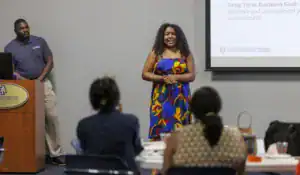 A woman in a colorful dress presents at a podium while a man listens. Audience members are seated at tables, watching the presentation. A screen displays information about business goals.