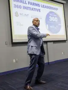A speaker gestures while presenting about the Small Farms Leadership 360 Initiative. A screen in the background displays the program's logo and information from North Carolina A&T State University.