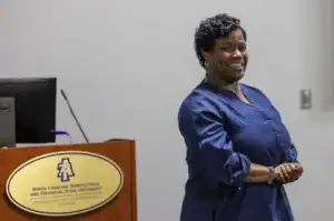 A woman with curly hair smiles while standing in front of a podium with a university logo. She appears engaged and is wearing a blue blouse.