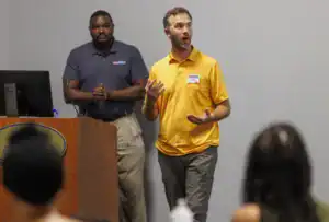 A man in an orange shirt speaks passionately while gesturing during a presentation, with a colleague in a dark shirt standing behind him at a podium. Attendees are seated, listening attentively.