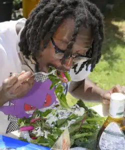 A person with dreadlocks eats a large salad, leaning in to take a bite. They wear glasses and a colorful shirt, surrounded by greens and a bottle of balsamic vinaigrette. The scene is outdoors with green grass in the background.