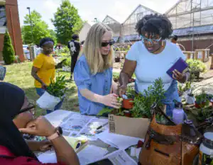 Participants engage in a gardening activity, examining plants and sharing tips. One person inspects a potted plant while another carries a bag of greenery. Outdoor setting with green trees and greenhouses in the background.