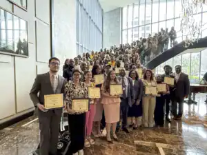 A large group of diverse individuals stands together in a modern venue, holding certificates. They pose on a staircase, with natural light streaming in through large windows.