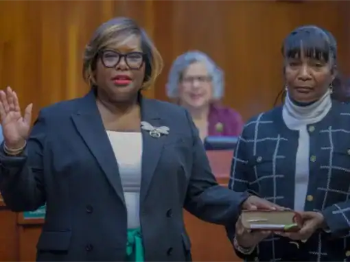 A woman is taking an oath with her right hand raised while holding a book, accompanied by another woman. A group is visible in the background, all in a formal setting.