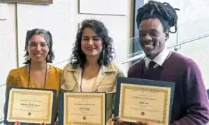 Three individuals stand together proudly holding their certificates of achievement, representing first, second, and third places in a competition. They are smiling and dressed in professional attire.