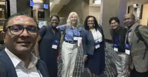 A diverse group of six people smiles together at a conference venue, wearing name badges and holding drinks. The setting features modern decor with screens in the background.