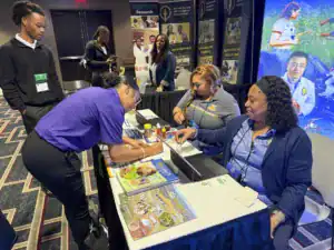 Three people at a conference booth are discussing materials. One person in a purple shirt is writing, while two others in blue shirts are seated at a table with laptops and brochures. A young man observes from behind.