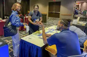 A young woman in a blue shirt holds a handout while engaging in conversation with a man at a table filled with brochures. Another woman observes from nearby, all in a conference setting.